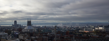 Liverpool Cathedrals wide This urban landscape photograph captures a wide view of Liverpool in the United Kingdom during a late autumn morning. The main subjects are the prominent churches: the Liverpool Metropolitan Cathedral with its distinctive modern architecture, and the traditional Liverpool Cathedral, both visible against the city skyline. Low clouds cover the sky, with hints of morning light breaking through, and smoke or mist can be seen rising in the distance. The River Mersey and the distant Welsh hills are visible in the background, adding depth to the scene. The image showcases the blend of historic and contemporary architecture that defines Liverpool, highlighting both the Liverpool Cathedral and Liverpool Metropolitan Cathedral as significant landmarks within this urban setting.